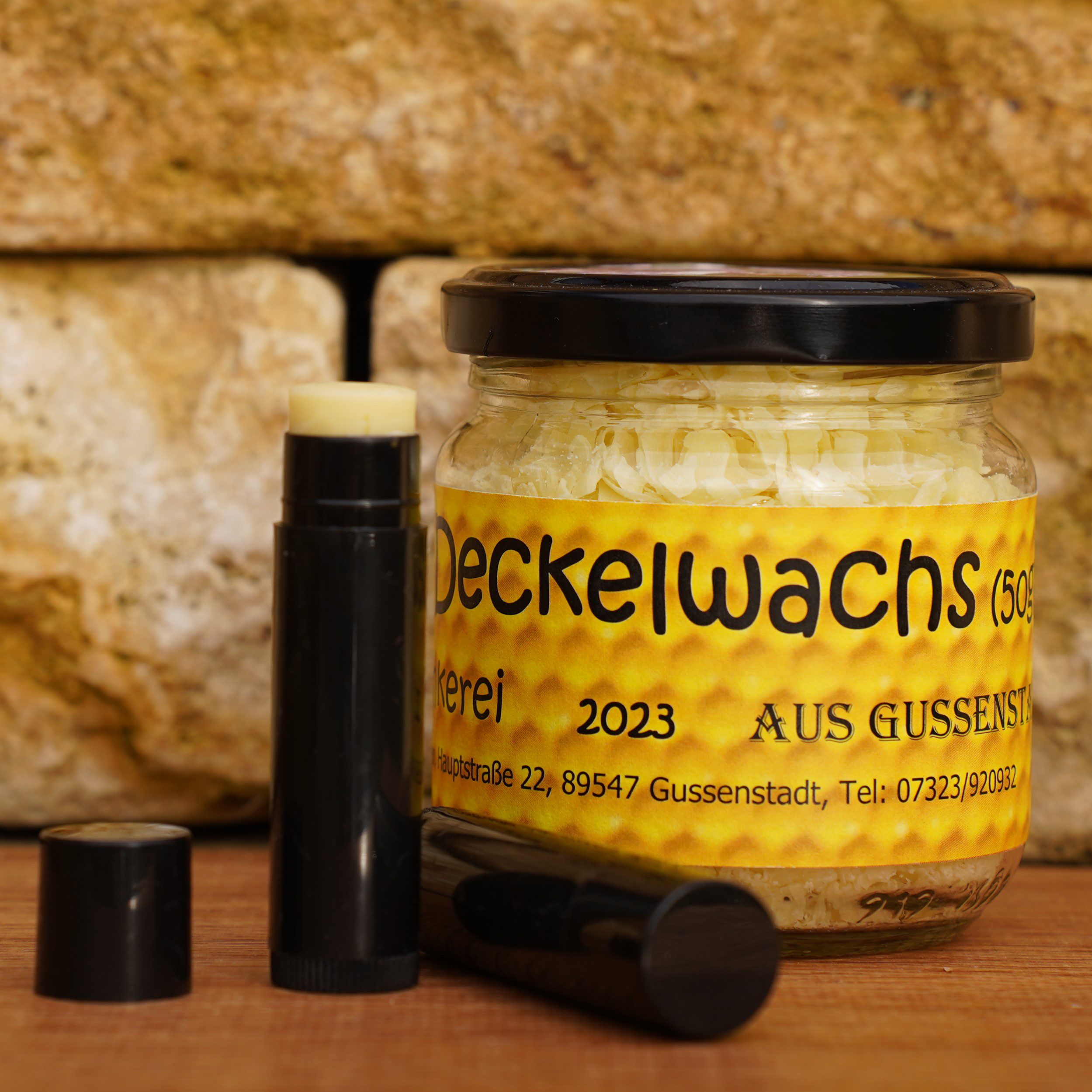 Ein Glas Deckelwachs mit schwarzem Deckel steht neben zwei schwarzen Lippenpflegestiften, vor einem Hintergrund aus beigen Ziegelsteinen.
