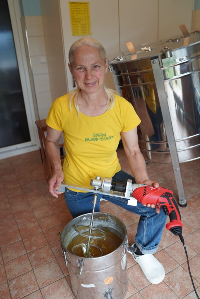 Frau mit gelbem T-Shirt rührt Honig mit einem elektrischen Rührgerät in einem Edelstahleimer.
